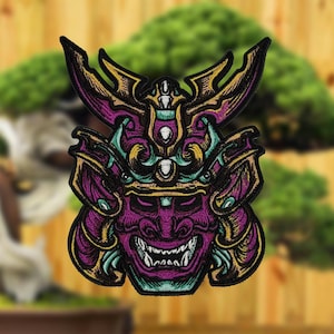May include: Embroidered patch featuring a purple, gold, and teal Oni mask design. The mask has a fierce expression with sharp teeth and intricate details. The patch is likely for jackets, bags, or other apparel.