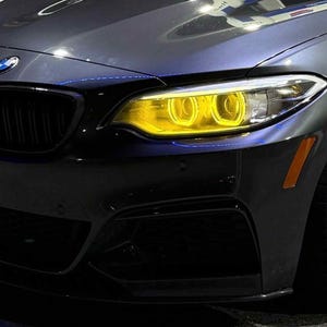 May include: Close-up of a dark gray BMW car with yellow headlights and a black grill. The car has a silver rim and a black front bumper. The car is parked on a gray surface.
