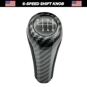May include: A black and gray 6-speed shift knob with a carbon fiber pattern. The shift pattern is displayed on the top of the knob with the letters R, 1, 3, 5, 2, 4, and 6. The knob has a rounded top and a textured grip.