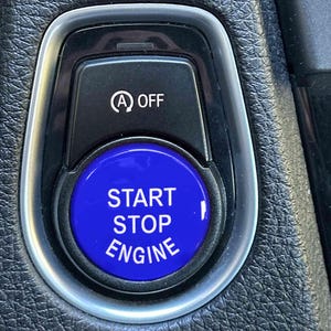 May include: Close-up of a car's dashboard featuring a black panel with a blue "START STOP ENGINE" button. Above the button is a smaller button with the "A OFF" symbol. The panel is surrounded by a silver trim.