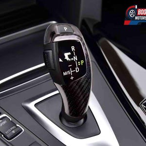 以下が含まれることがあります： カーボンファイバーパターンの車のギアシフトレバーのクローズアップ。レバーには、P、R、N、D、M/Sのギアポジションが表示されています。「SPORT」と「ECO PRO」ボタンも表示されています。