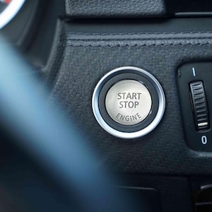 May include: Close-up of a car's dashboard featuring a silver start/stop engine button with the text "START STOP ENGINE". The button is surrounded by a silver ring and set against a textured black surface. An air vent is visible to the right.