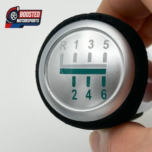 Pode incluir: Um botão de mudança de marcha prateado com uma pega preta. O botão apresenta um padrão de engrenagens com os números R, 1, 3, 5, 2, 4 e 6 em verde. O logótipo "Boosted Motorsports" é visível no canto superior esquerdo.