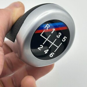 Puede incluir: Un pomo de palanca de cambios plateado y negro con un agarre de cuero negro. El pomo presenta una cara negra con números de marcha blancos y un diseño de rayas rojas, azules y negras. El patrón de cambio de marchas es visible.