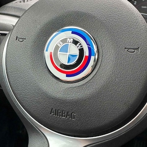 Puede incluir: Primer plano de un volante negro con un emblema de BMW. El emblema es circular con detalles en azul, blanco, rojo y plateado. La palabra "AIRBAG" está impresa en blanco en el volante.