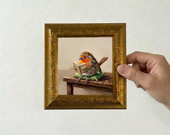 May include: A square painting in an ornate gold frame depicting a small robin with an orange breast perched on a green cushion atop a wooden table. The bird is holding an open book with its wings, appearing to read while resting on the surface.