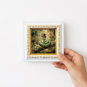May include: A framed painting of a green snake wearing a brown top hat, perched on a tree branch. The artwork is set against a lush, green forest background, and the frame is white with gold accents.