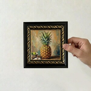 May include: A square oil painting in a black and gold ornate frame depicting a pineapple centered on a wooden surface with a small bowl of green limes to the left. The background features blended warm tones of orange, yellow, and brown paint.