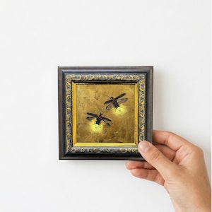 May include: A small square painting in a dark ornate frame with gold accents. The artwork depicts two fireflies with glowing yellow abdomens against a textured, metallic gold background, capturing the insects in flight with detailed wings.