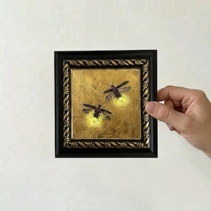May include: A square painting in a black frame with a decorative rope-like border. The artwork features two fireflies with dark bodies and translucent wings against a textured gold background, each emitting a soft, glowing yellow light.
