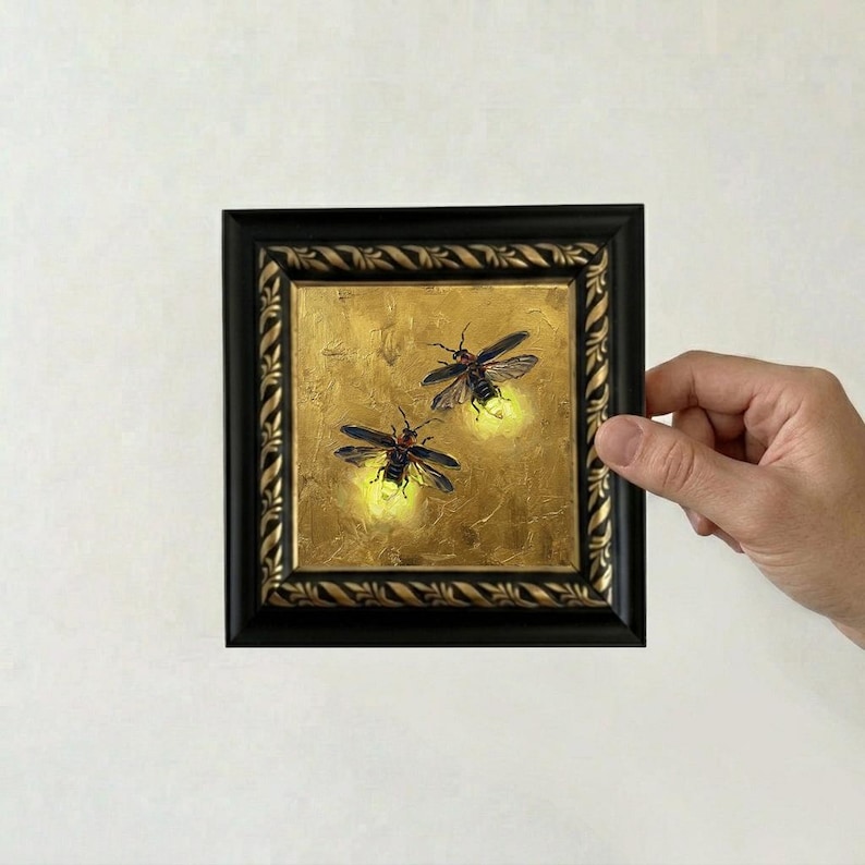 May include: A square painting in a black frame with a decorative rope-like border. The artwork features two fireflies with dark bodies and translucent wings against a textured gold background, each emitting a soft, glowing yellow light.