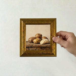 May include: A square oil painting in a decorative gold frame depicting a still life of several brown potatoes on a rustic wooden surface. One potato is sliced into thin rounds, revealing a light cream-colored interior against a neutral background.