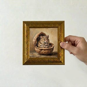 May include: A square oil painting in an ornate gold frame depicting a small brown mouse sitting inside a cracked walnut shell. The mouse has light brown fur, dark eyes, and is positioned centrally within the shell against a neutral background.