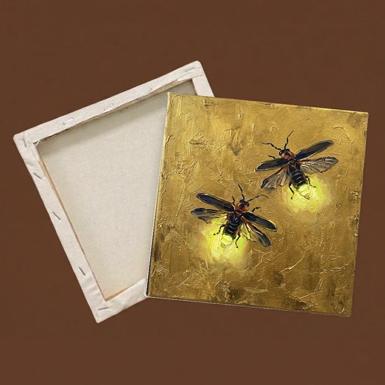 May include: A square canvas painting featuring two fireflies with glowing yellow abdomens against a textured metallic gold background, shown alongside an empty wooden stretcher frame with a plain white canvas back.
