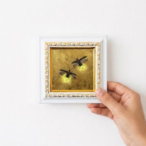 May include: A small square painting of two fireflies with glowing yellow abdomens against a textured gold background, encased in a white frame with ornate gold leaf detailing along the inner and outer edges of the decorative border.