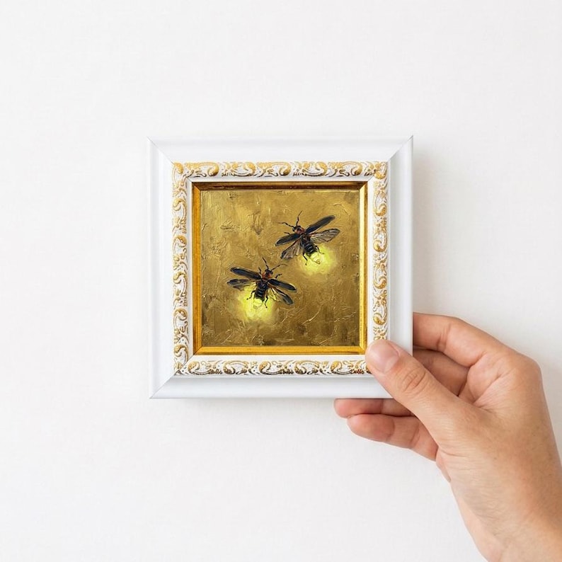 May include: A small square painting of two fireflies with glowing yellow abdomens against a textured gold background, encased in a white frame with ornate gold leaf detailing along the inner and outer edges of the decorative border.