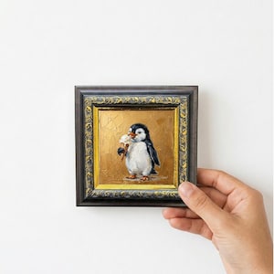 May include: A small square oil painting in a dark, ornate frame with gold accents. The artwork depicts a black and white penguin holding a light-colored ice cream cone against a textured, metallic gold background, rendered with visible brushstrokes.