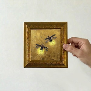 May include: A square painting in an ornate gold frame featuring two fireflies with glowing yellow abdomens against a textured gold background. The insects have dark wings and bodies, with bright light emanating from their lower sections.