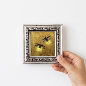 May include: A small square painting in an ornate silver frame featuring two fireflies with glowing yellow abdomens against a textured gold background. The artwork depicts the insects in flight, capturing the bioluminescent light they emit.
