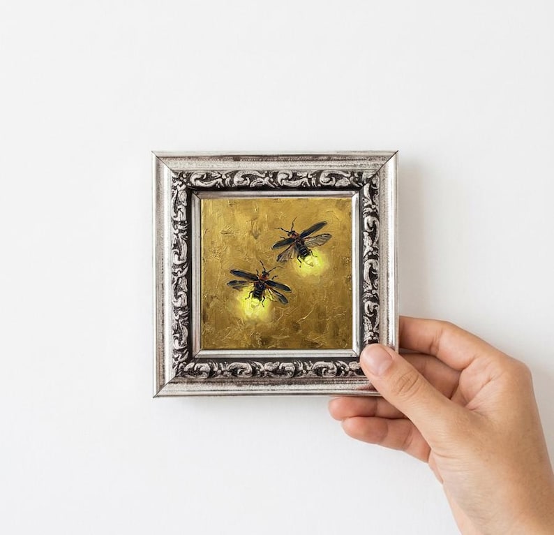 May include: A small square painting in an ornate silver frame featuring two fireflies with glowing yellow abdomens against a textured gold background. The artwork depicts the insects in flight, capturing the bioluminescent light they emit.