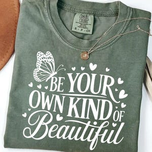 Puede incluir: Camiseta verde oliva con la frase "BE YOUR OWN KIND OF BEAUTIFUL" en escritura blanca y un gráfico de mariposa. También se ven un collar dorado y un sombrero marrón.