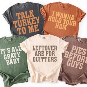 Peut inclure: Cinq t-shirts à manches courtes de différentes couleurs, chacun avec une phrase humoristique sur le thème de Thanksgiving en lettres grasses et usées. Les chemises sont dans des tons de gris, orange, vert, crème et marron. Les phrases incluent "Talk Turkey To Me" et "I Wanna Hold Your Ham."