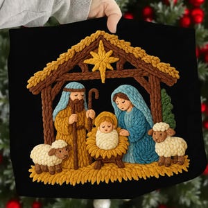 May include: Black Christmas-themed pillow featuring a detailed embroidered nativity scene. The scene includes Mary, Joseph, baby Jesus, sheep, and a star, all set within a rustic stable. The embroidery uses shades of brown, blue, yellow, and white.