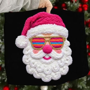 May include: A black pillow featuring a crocheted Santa Claus face. Santa wears a pink hat, gold-framed sunglasses with rainbow lenses, and has a pink nose and smiling mouth. The beard is white and fluffy.