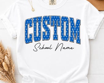May include: A white short-sleeved t-shirt featuring the word CUSTOM in large blue block letters with a white polka dot pattern. Below the word CUSTOM, the text School Name is printed in a black cursive script. The shirt has a crew neckline.