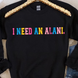 Op de afbeelding: Zwarte crewneck sweatshirt met de tekst "I NEED AN ALANI" in kleurrijke, vervaagde letters. De sweatshirt wordt getoond op een gouden paillettenhanger, met een spijkerbroek en bruine laarzen op de achtergrond.