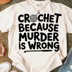 May include: Cream-colored t-shirt with the text "CROCHET BECAUSE MURDER IS WRONG" in bold black letters. A yarn ball and crochet hook graphic are above the word "CROCHET". A crochet hook graphic is below the text.