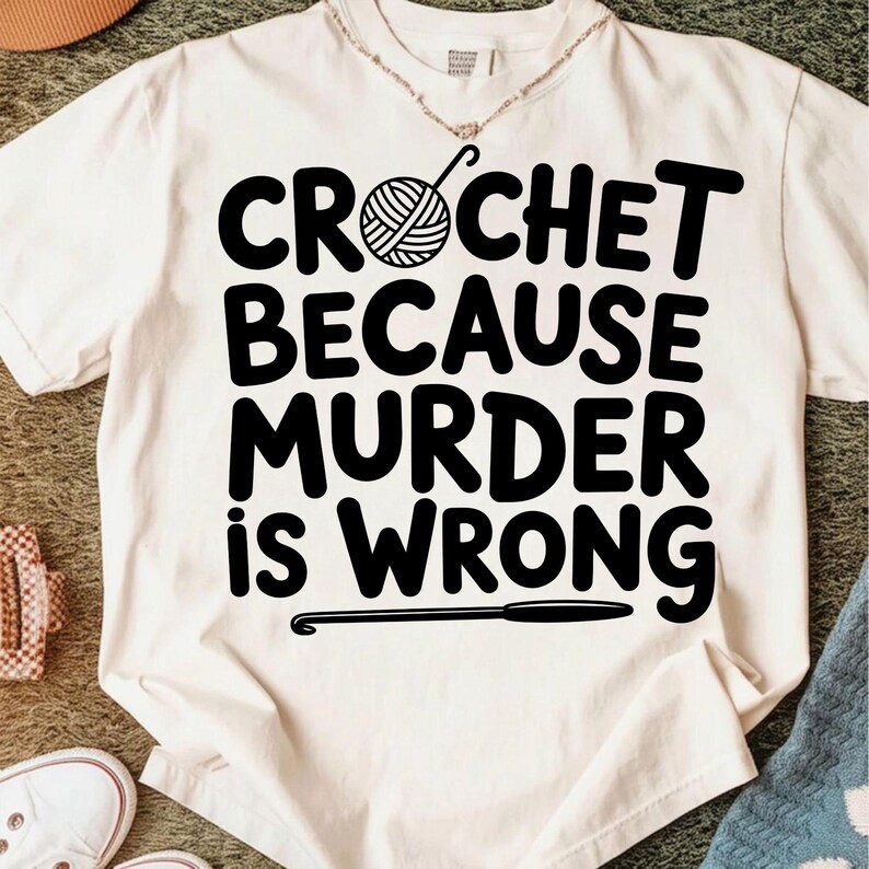 May include: Cream-colored t-shirt with the text "CROCHET BECAUSE MURDER IS WRONG" in bold black letters. A yarn ball and crochet hook graphic are above the word "CROCHET". A crochet hook graphic is below the text.
