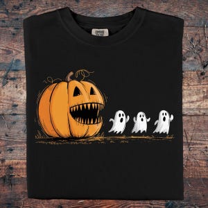 Puede incluir: Camiseta negra con un diseño de Halloween. Una calabaza naranja con una gran sonrisa dentada es seguida por tres fantasmas blancos de dibujos animados. El diseño está impreso en una camiseta negra Comfort Colors.