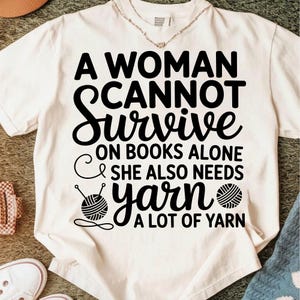 May include: Cream-colored t-shirt with the text "A Woman Cannot Survive on Books Alone & She Also Needs Yarn A Lot of Yarn" in black. The design includes a yarn ball and knitting needles.