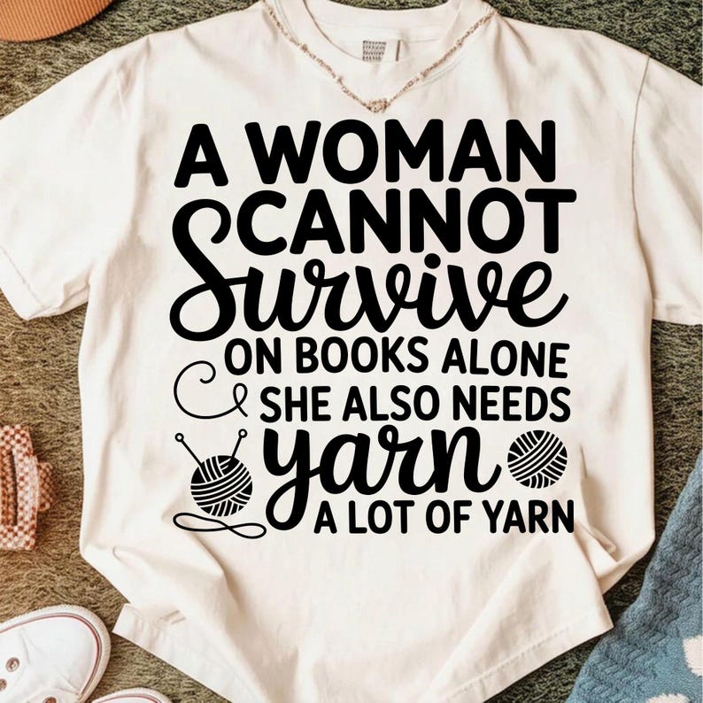 May include: Cream-colored t-shirt with the text "A Woman Cannot Survive on Books Alone & She Also Needs Yarn A Lot of Yarn" in black. The design includes a yarn ball and knitting needles.