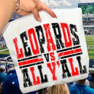 May include: White pillow with the text "LEOPARDS VS ALL Y'ALL" in red and black block letters. The pillow is being held up in front of a football stadium.