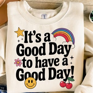 K&ouml;nnte beinhalten: Ein cremefarbener Pullover mit dem Text "It's a Good Day to have a Good Day!" in Schwarz. Der Text ist umgeben von einem Regenbogen, einem gelben Stern, einer rosa Blume, einem Smiley und zwei roten Kirschen.
