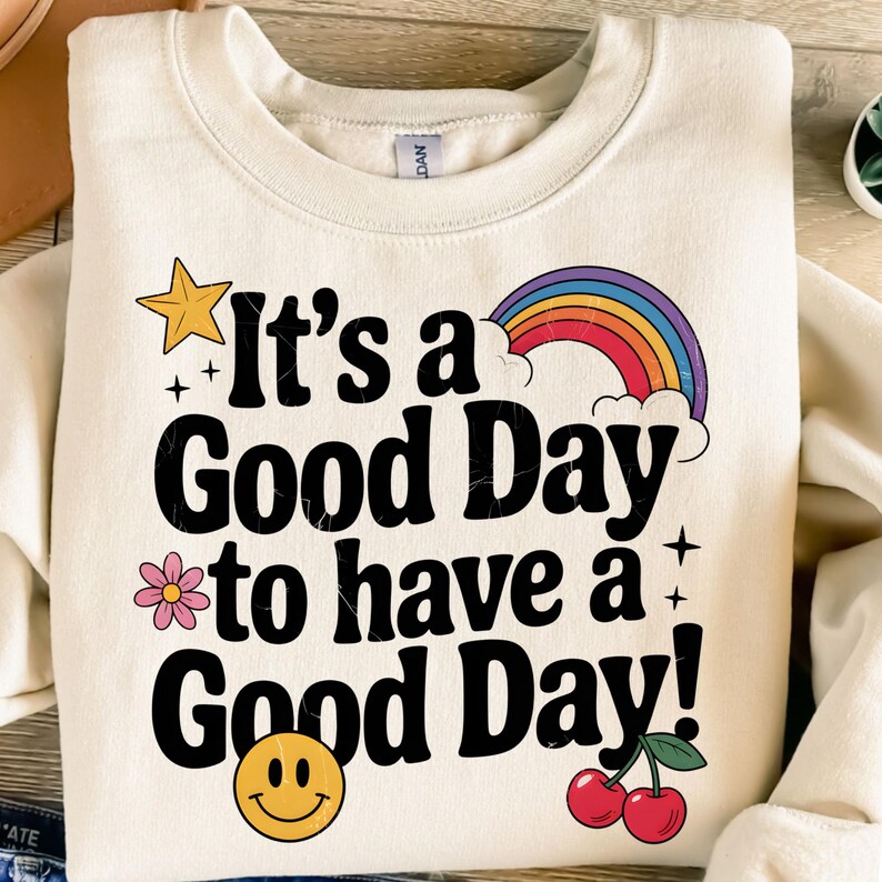 K&ouml;nnte beinhalten: Ein cremefarbener Pullover mit dem Text "It's a Good Day to have a Good Day!" in Schwarz. Der Text ist umgeben von einem Regenbogen, einem gelben Stern, einer rosa Blume, einem Smiley und zwei roten Kirschen.