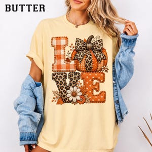 Puede incluir: Una camiseta de manga corta de color amarillo pálido con un estampado gráfico que forma la palabra LOVE. La L es de cuadros naranjas, la O es una calabaza con un lazo de leopardo, la V es de estampado de leopardo y la E es naranja con lunares blancos.