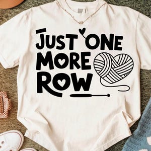May include: Cream-colored t-shirt with the text "JUST ONE MORE ROW" in bold black letters. A heart shape made of yarn and a crochet hook are also featured in black. The shirt is a casual garment.