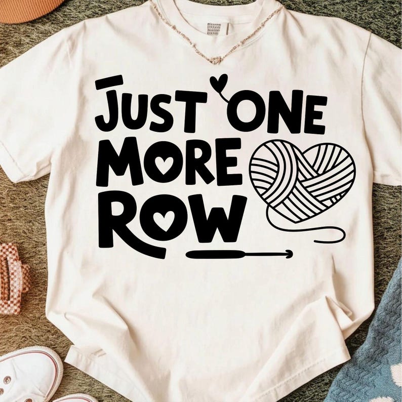 May include: Cream-colored t-shirt with the text "JUST ONE MORE ROW" in bold black letters. A heart shape made of yarn and a crochet hook are also featured in black. The shirt is a casual garment.