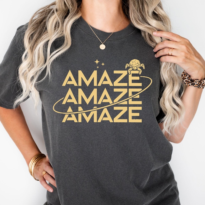 Op de afbeelding: Donkergrijs t-shirt met het woord "AMAZE" in goud, drie keer gestapeld met een planeet-afbeelding en een klein wezen. Het t-shirt heeft korte mouwen en een ronde hals.