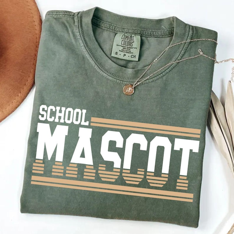 May include: An olive green t-shirt with the words "SCHOOL MASCOT" in white block letters and tan horizontal stripes. A silver necklace with a small pendant rests on the shirt. The shirt is folded and placed on a white surface.