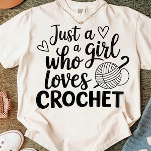 May include: Cream-colored t-shirt with the text "Just a girl who loves CROCHET" in black script. The design includes a heart and a yarn ball with a crochet hook. The shirt is a casual wear item.