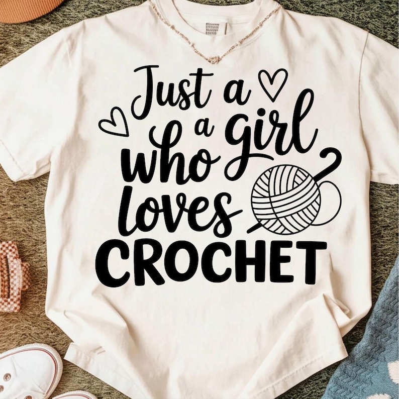 May include: Cream-colored t-shirt with the text "Just a girl who loves CROCHET" in black script. The design includes a heart and a yarn ball with a crochet hook. The shirt is a casual wear item.