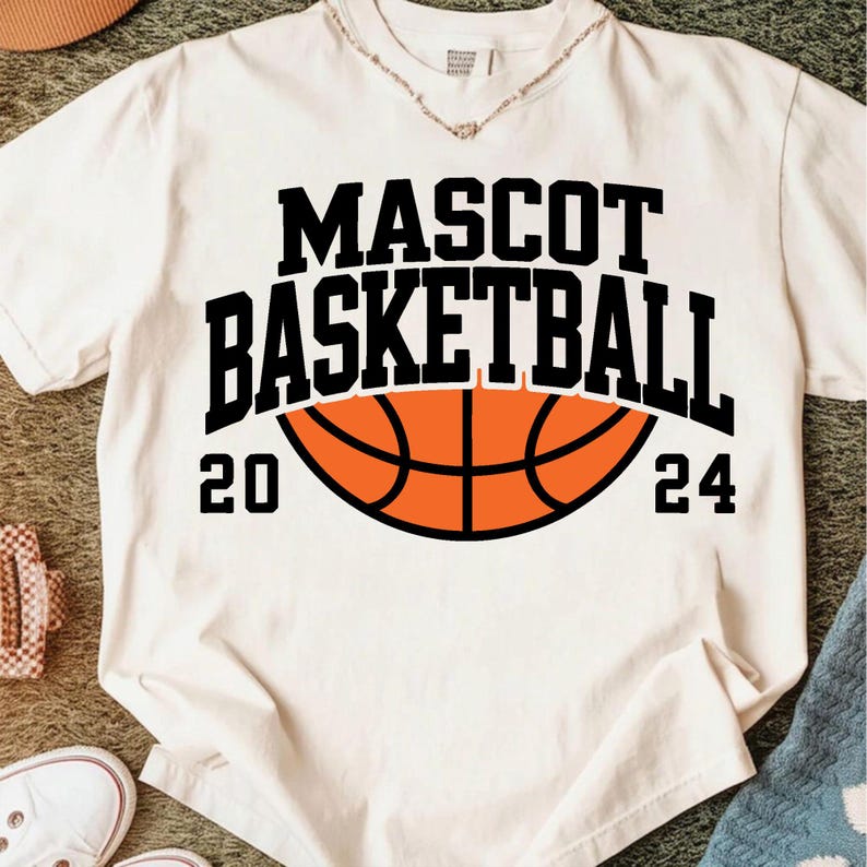 Peut inclure: T-shirt blanc cass&eacute; avec l'inscription noire "MASCOT BASKETBALL" au-dessus d'un ballon de basket orange. Les chiffres "20" et "24" encadrent le ballon. V&ecirc;tement d&eacute;contract&eacute;.