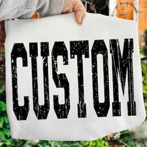 May include: A white fabric item with the word "CUSTOM" printed in large, distressed black letters. The text has a bold, collegiate-style font. The item is held up by a person, with a blurred outdoor background.