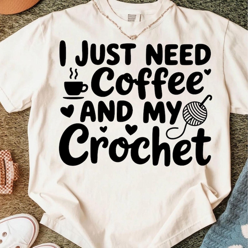 May include: Cream-colored t-shirt with the text "I JUST NEED Coffee AND MY Crochet" in black. A coffee cup, hearts, and a ball of yarn with a crochet hook are also featured in black. The shirt has a round neckline and short sleeves.