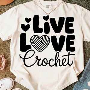 May include: Cream-colored t-shirt with the words "Live Love Crochet" in black. The word "Love" features a heart-shaped design with a yarn-like pattern. Two small hearts are above the word "Live".