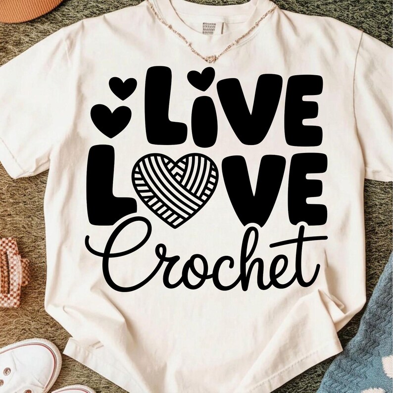 May include: Cream-colored t-shirt with the words "Live Love Crochet" in black. The word "Love" features a heart-shaped design with a yarn-like pattern. Two small hearts are above the word "Live".
